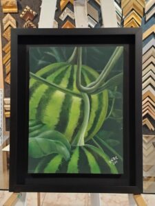 Pintura hiperrealista de una sandía entre hojas verdes, enmarcada con moldura negra tipo caja por Sol y Sombra SRL.