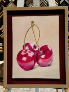 Pintura hiperrealista de tres cerezas rojas, enmarcada con moldura clara y fondo rojo por Sol y Sombra SRL.