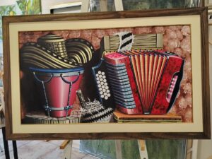 Pintura folclórica con acordeón, tambor y sombrero, enmarcada con moldura de madera por Sol y Sombra SRL.