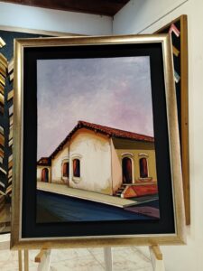 Pintura de casa colonial con techo de tejas, enmarcada con moldura dorada clara por Sol y Sombra SRL.