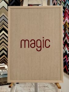 Cuadro con fondo de tela rayada en tonos beige y la palabra "magic" bordada en rojo, enmarcado por Sol y Sombra SRL.