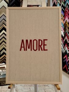 Cuadro decorativo con fondo de tela rayada en beige y la palabra "AMORE" bordada en rojo, enmarcado por Sol y Sombra SRL.