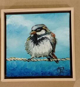Pintura de un pajarito posado sobre una cuerda con fondo azul, enmarcada con moldura clara por Sol y Sombra SRL.